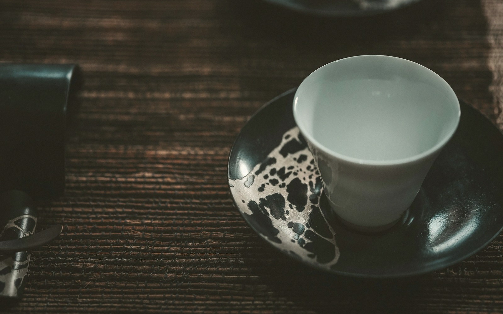 A white teacup rests on a dark speckled saucer atop a woven mat